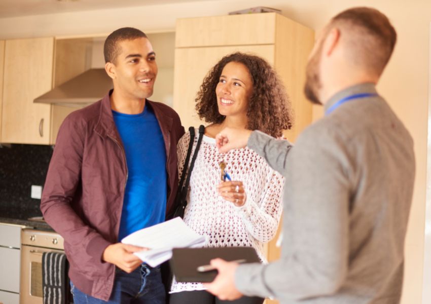 A landlord in a gray sweater hands the keys to the property to two smiling renters who are hugging at the side.